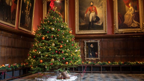 Christmas tree decorated with red, gold and orange baubles, with small lights and topped with a red box. There is a large portrait paintings on the walls either side of the tree. There is wooden panelling below these paintings, a wooden bench with a green foliage garland laid along it, and a chequered stone floor.